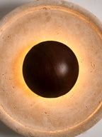 Close, lit view of this Clérachic wall light, with warm light spilling around the walnut dome onto the porous travertine face.