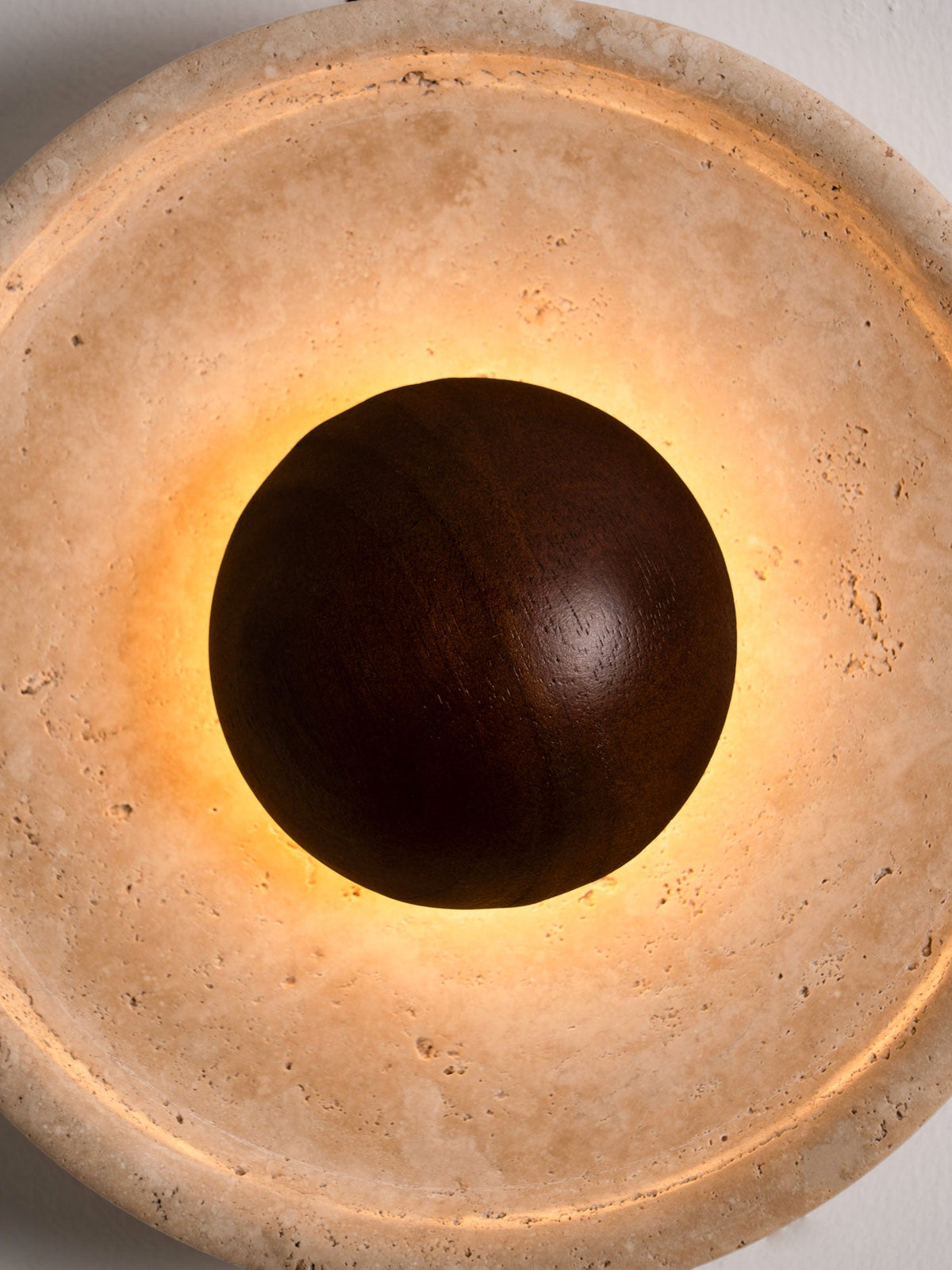 Close, lit view of this Clérachic wall light, with warm light spilling around the walnut dome onto the porous travertine face.