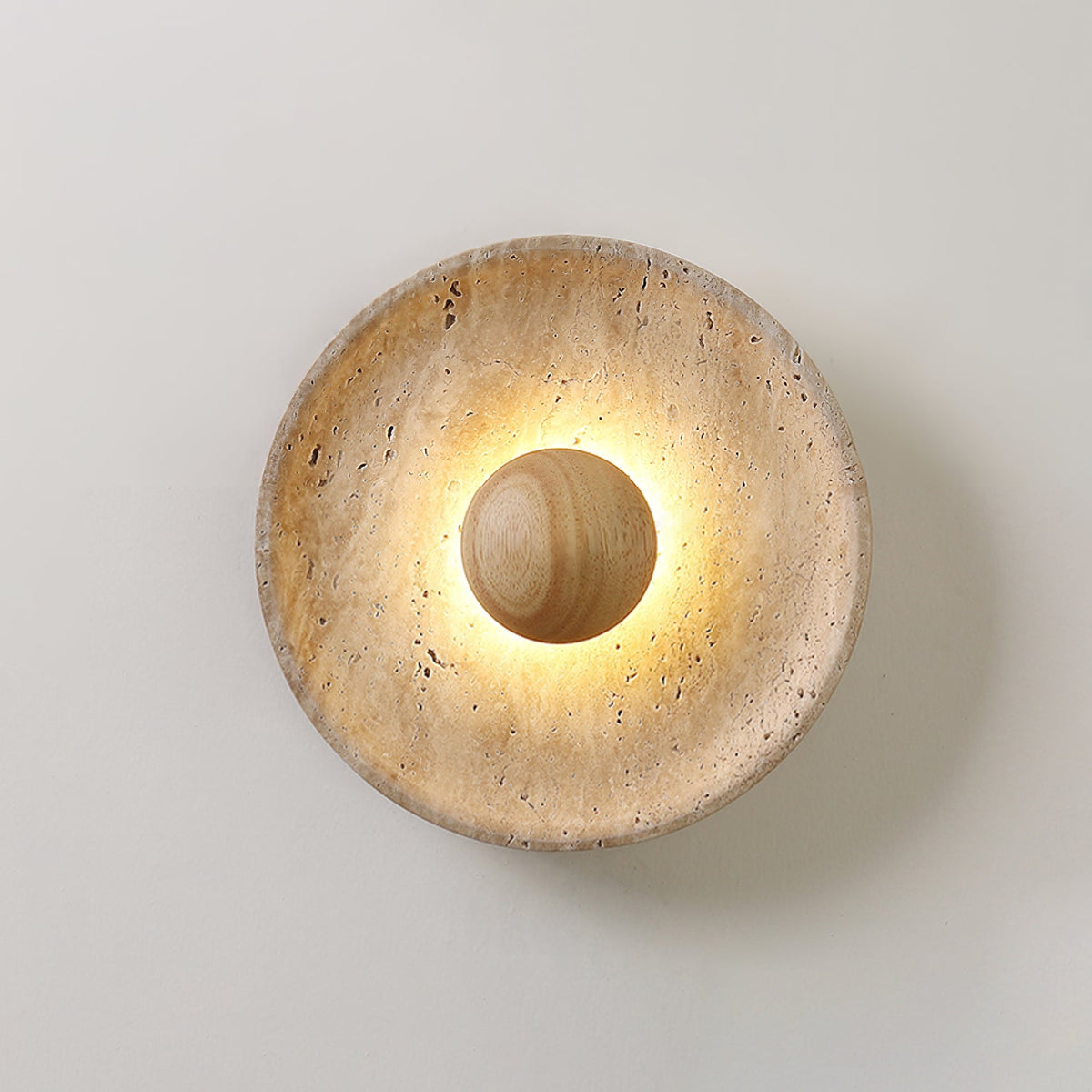 Clérachic travertine wall light lit warm, with a light wood dome at its centre and a glow around its pitted stone dish.