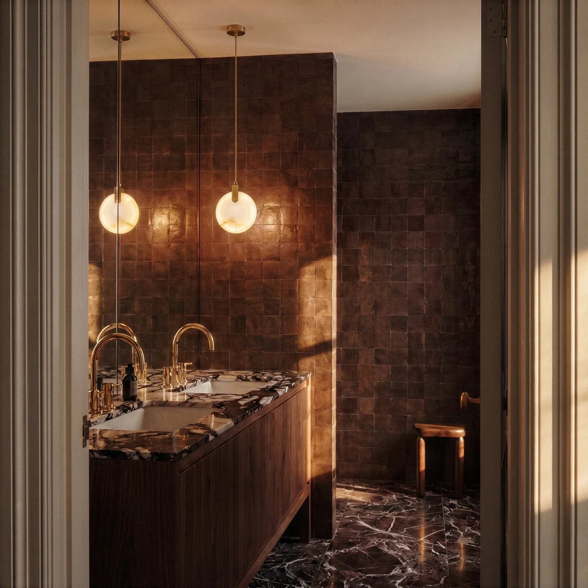 Clérachic pair of lit alabaster pendants on brass-toned cords above a double vanity in a dark tiled bathroom.
