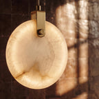 Clérachic lit alabaster pendant close up, its brass-toned bracket and glowing veined stone against dark glazed tiles.