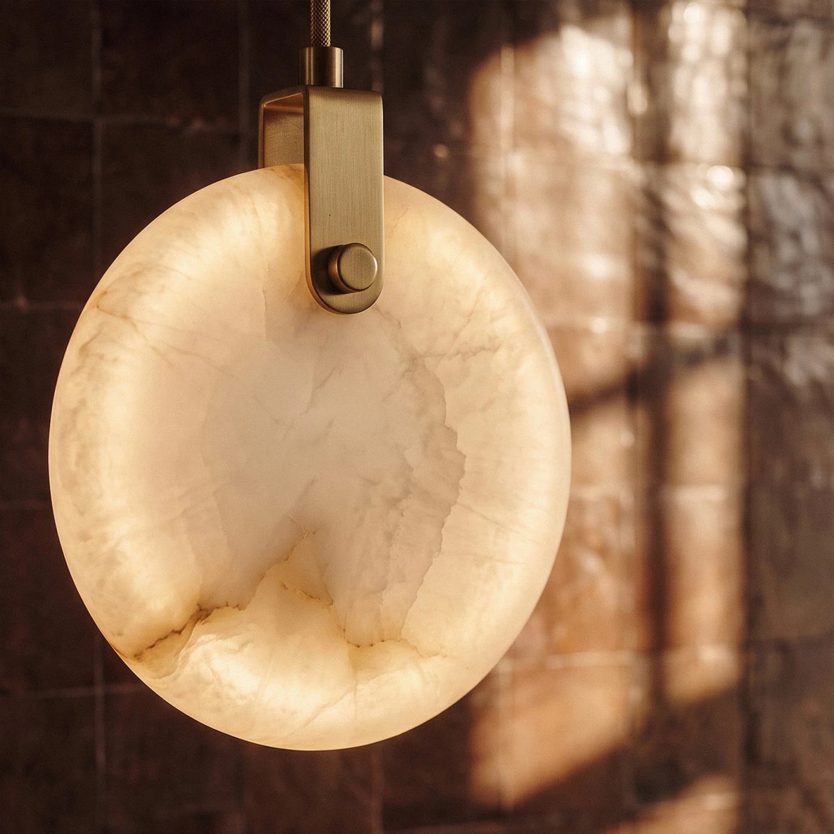 Clérachic lit alabaster pendant close up, its brass-toned bracket and glowing veined stone against dark glazed tiles.