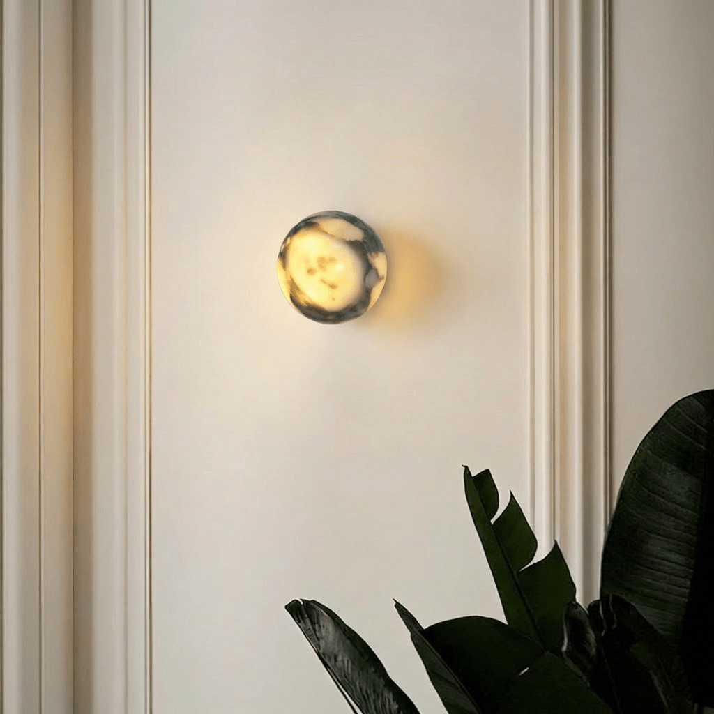 Clérachic wall lamp lit on a white panelled wall, spilling a soft halo of light behind it beside a large leafy houseplant.