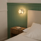 Clérachic wall lamp glowing on a green beadboard wall above a wooden nightstand and a cream upholstered headboard.