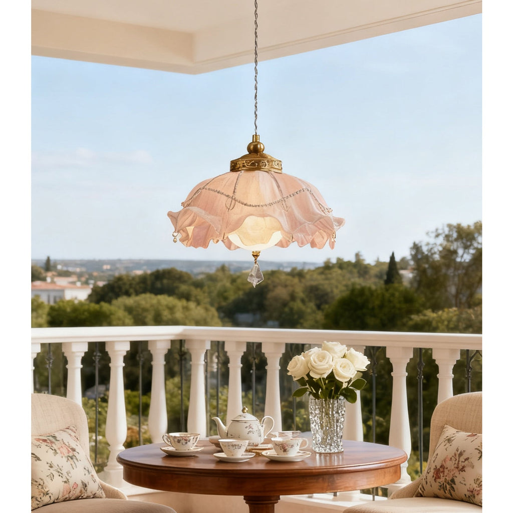 Pink-edged ruffled pendant from Clérachic hanging on a balcony above a round wooden table set with teacups, with trees beyond the railing.