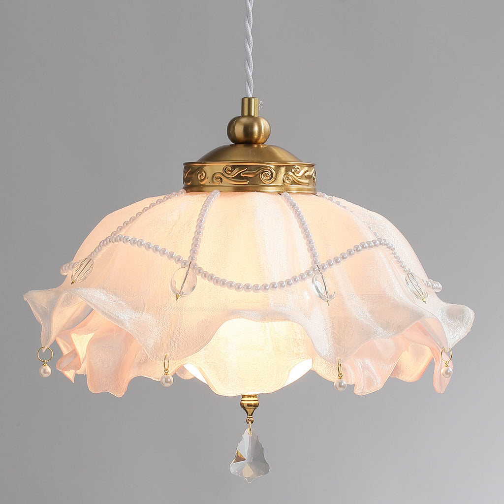 Glowing Clérachic pendant on a grey backdrop, its ruffled shade lit pale pink under a carved brass collar with draped pearl strands.