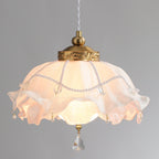 Glowing Clérachic pendant on a grey backdrop, its ruffled shade lit pale pink under a carved brass collar with draped pearl strands.