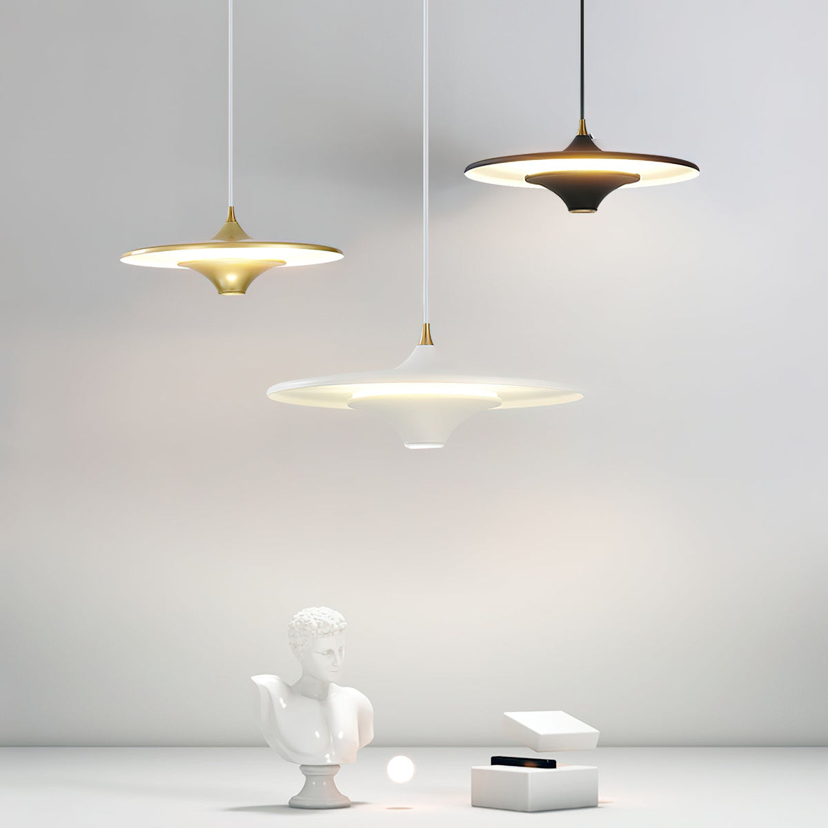 Three Clérachic saucer pendants in gold, black and white hang over a plaster bust and a small white box.