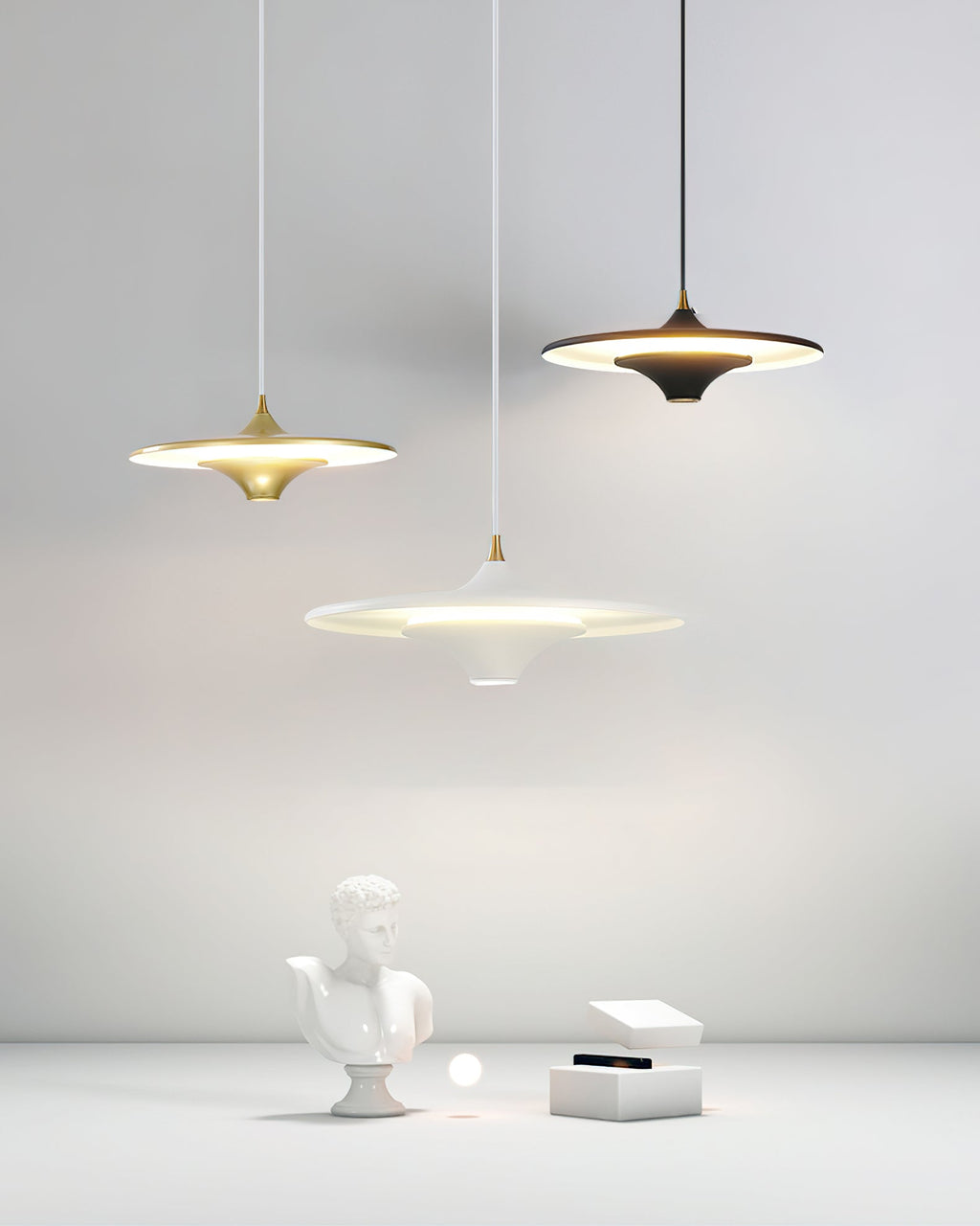 Three Clérachic saucer pendants in gold, black and white are lit above a plaster bust and white box in a light room.