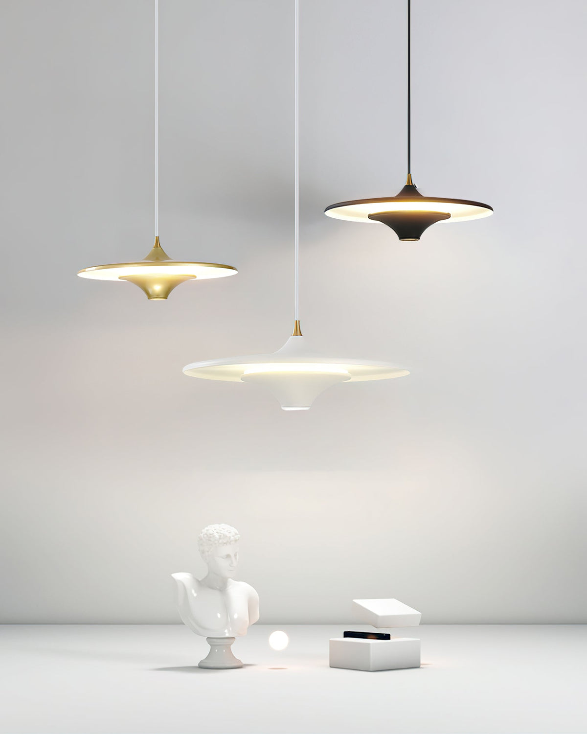 Three Clérachic saucer pendants in gold, black and white are lit above a plaster bust and white box in a light room.