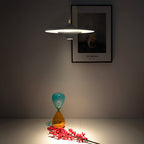 A Clérachic saucer pendant casts a pool of light in a dim room, with a framed print behind and a glass hourglass below.