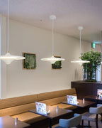 Three white Clérachic saucer pendants hang in a café above leather banquette seating, with framed greenery on the wall.