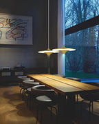 Two Clérachic saucer pendants with gold collars hang over a long timber table in a dark room with tall windows.