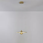Gold Clérachic saucer pendant with a brass canopy and a warm glow, hanging against a pale grey wall.