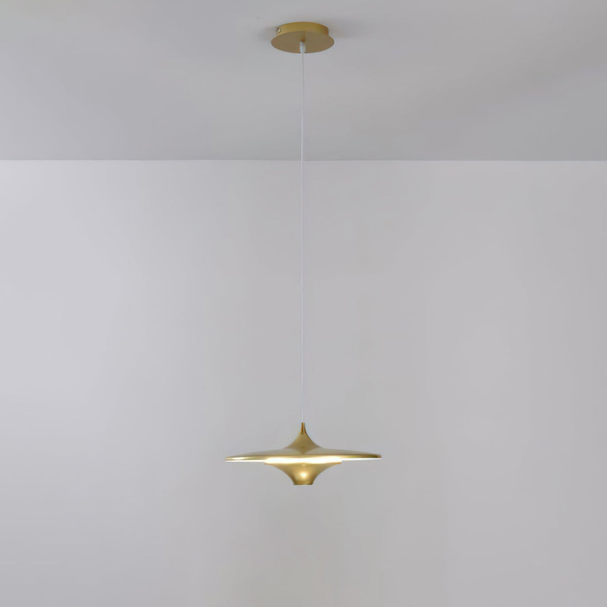 Gold Clérachic saucer pendant with a brass canopy and a warm glow, hanging against a pale grey wall.