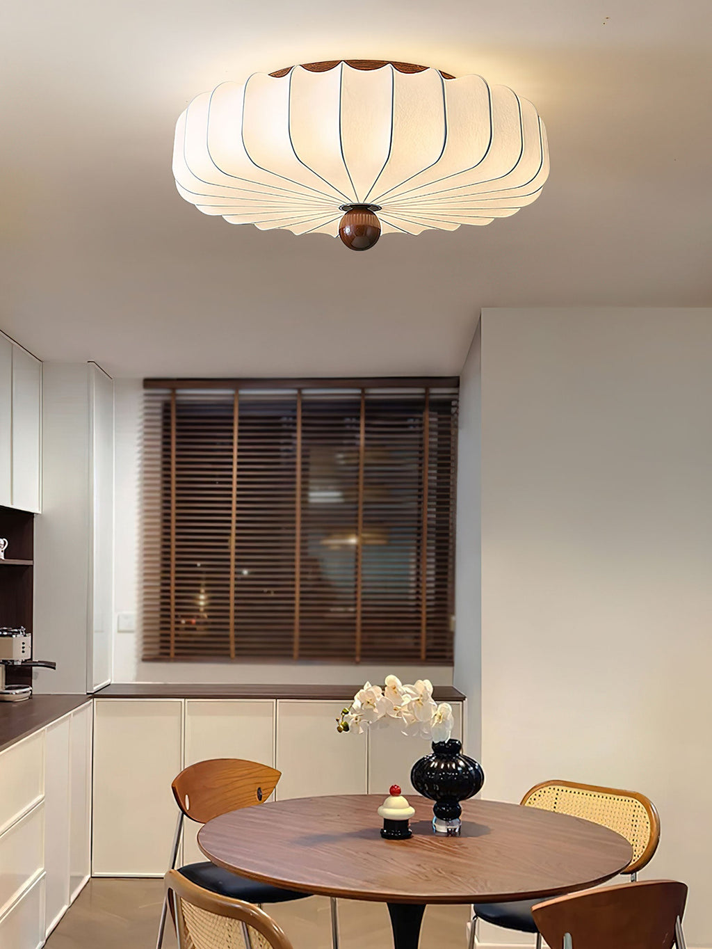 Above a kitchen dining corner, this Clérachic ceiling light with a pleated white shade hangs over a round wooden table and cane chairs.