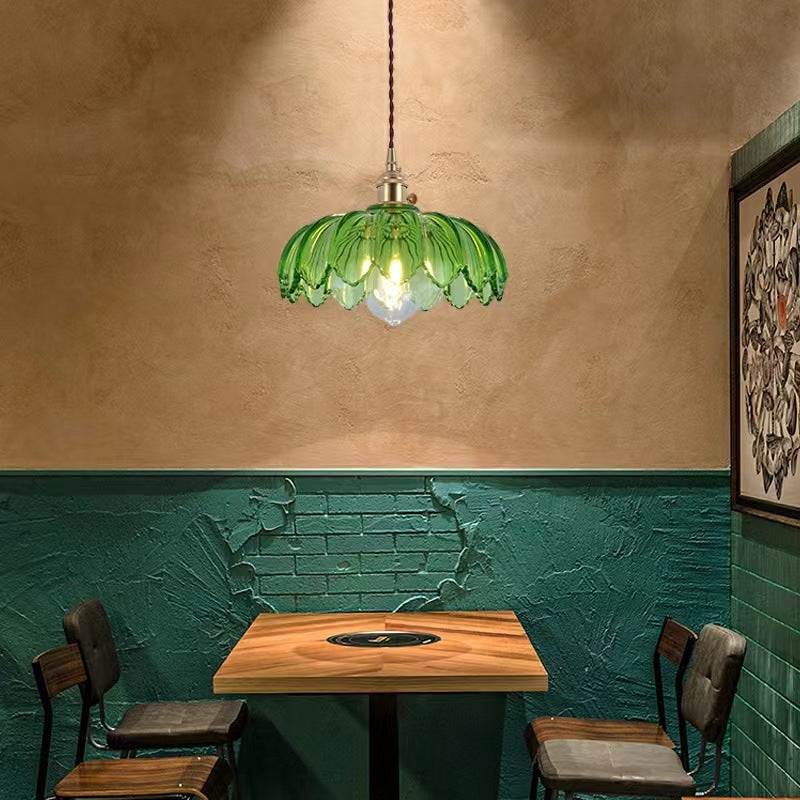 The Clérachic Scalloped Flower Glass Pendant Light in green glass above a small wooden café table set against a teal brick wall.