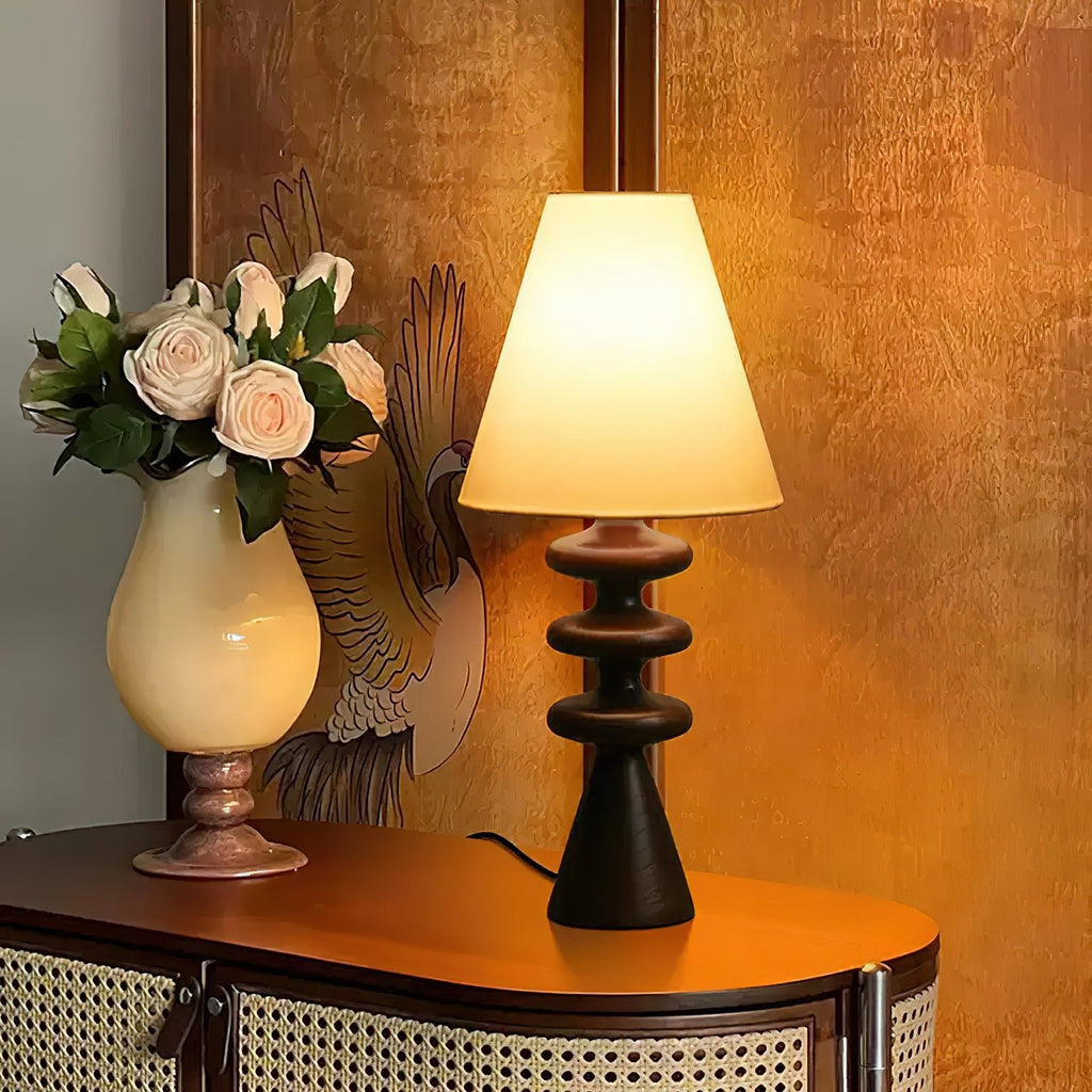 Black-based Clérachic table lamp glowing on a curved cane-fronted cabinet, beside a cream vase of roses against an amber wall.