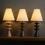 Three Clérachic table lamps in a row on a dark shelf, their rippled bases in wood tone, walnut and black, all with lit white shades.