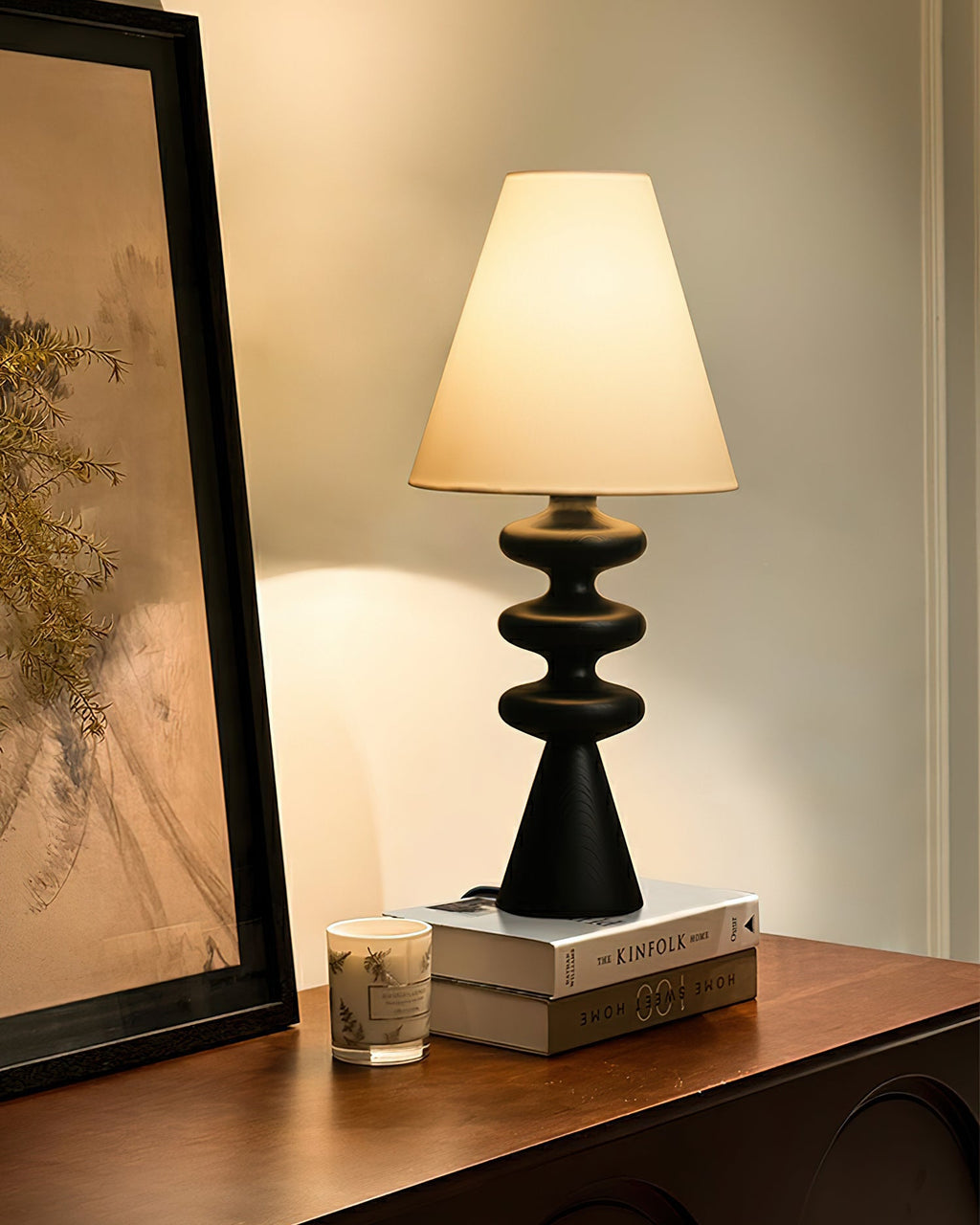 Black Clérachic table lamp on a wooden sideboard beside a leaning framed picture, a stack of books and a small candle.