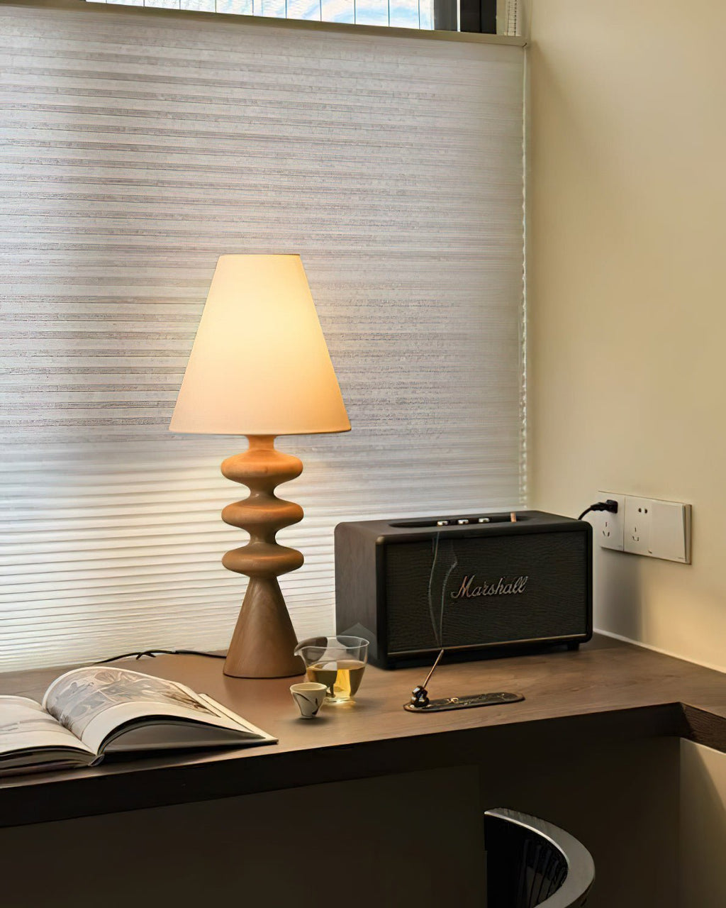 Wood-tone Clérachic table lamp lit on a corner desk by a window with a pleated blind, next to an open magazine and a speaker.