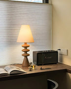 Wood-tone Clérachic table lamp lit on a corner desk by a window with a pleated blind, next to an open magazine and a speaker.