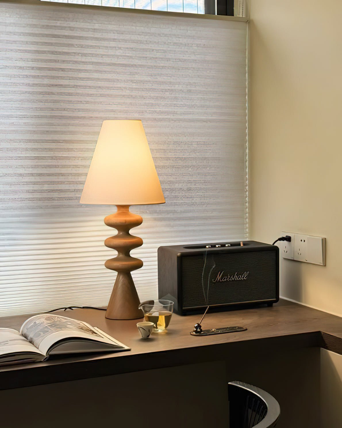 Wood-tone Clérachic table lamp lit on a corner desk by a window with a pleated blind, next to an open magazine and a speaker.