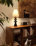 Small black Clérachic table lamp lit on a wooden bookshelf, a leafy plant to one side and a grey cat looking up below.