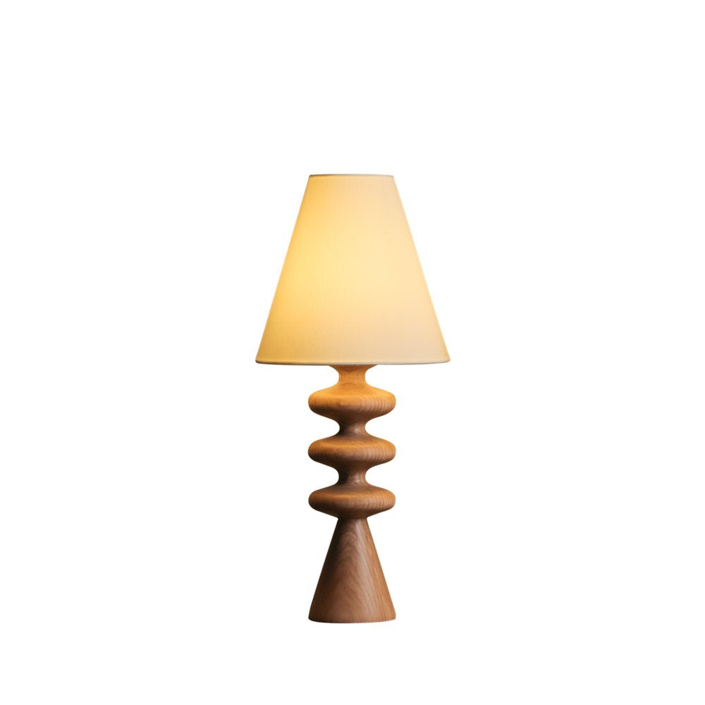 Wood-tone Clérachic table lamp with a lit white fabric shade and a rippled turned base, on a plain white backdrop.