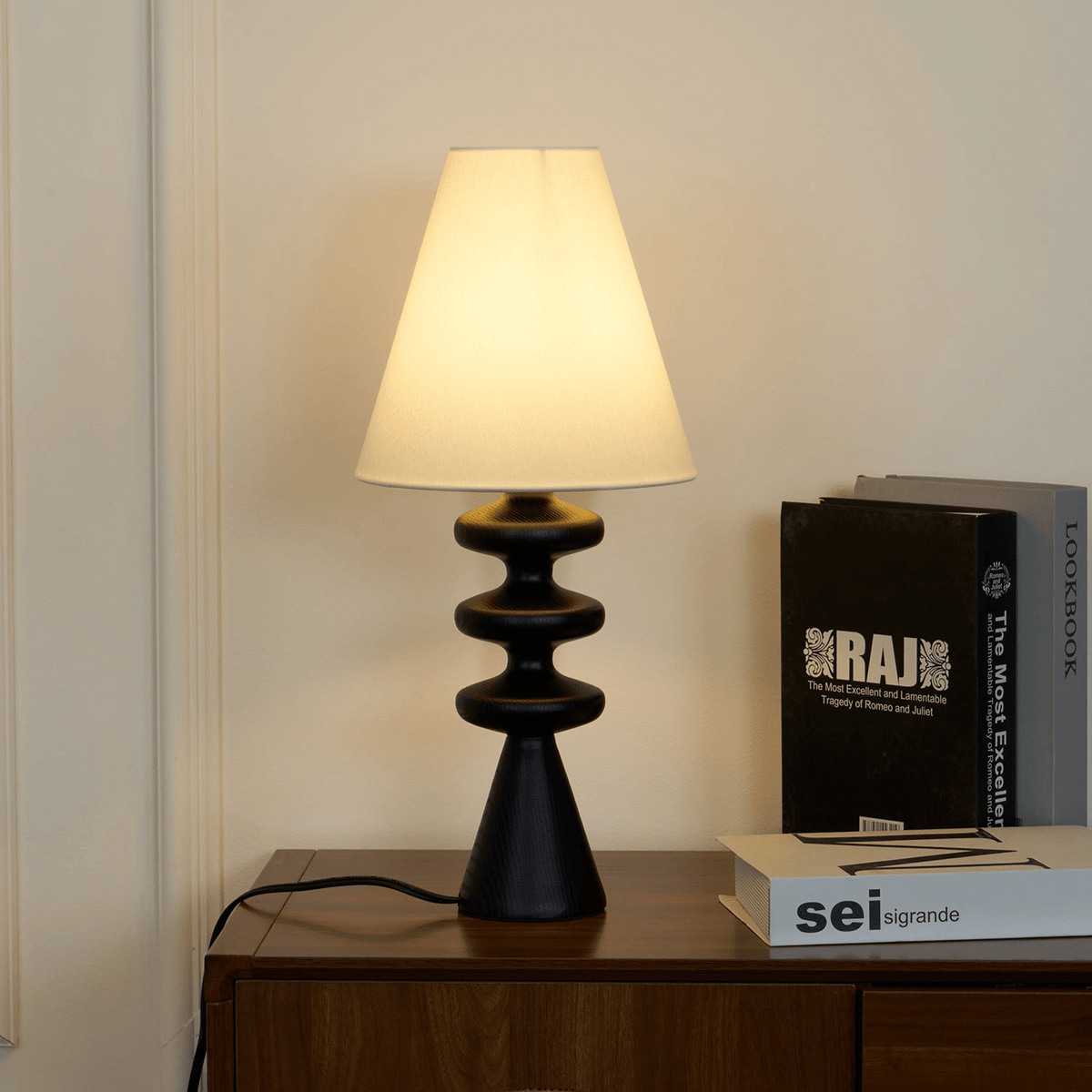 Lit Clérachic table lamp seen from a lower angle on a walnut cabinet, its cord trailing behind a pile of art books.