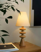 Wood-tone Clérachic table lamp glowing beside a trailing plant on a wooden surface, with a magazine at its foot.