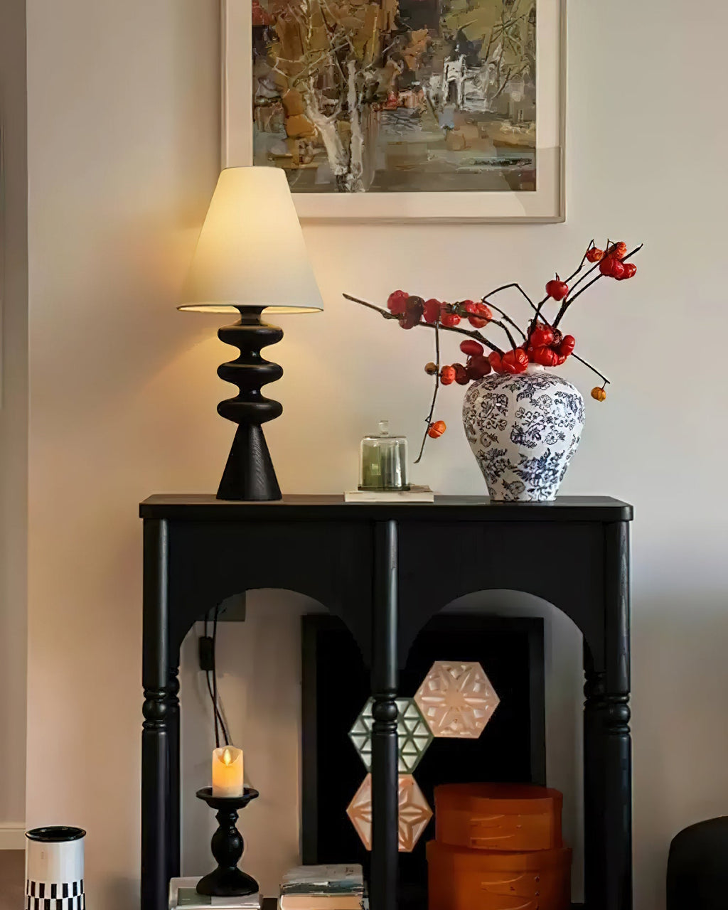 Black Clérachic table lamp on a black arched console, with a blue-and-white vase of red berries and a framed landscape above.