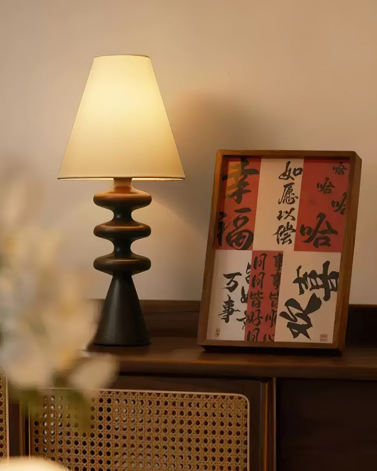 Black Clérachic table lamp on a wooden sideboard beside a framed red and cream calligraphy print, cane panelling below.