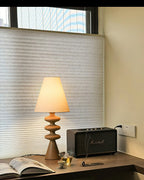 Wood-tone Clérachic table lamp on a desk by a window with a pleated blind, an open magazine and a speaker nearby.