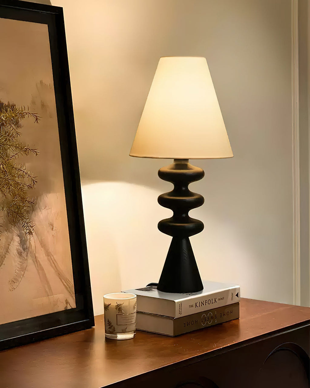 Close view of a black Clérachic table lamp glowing beside a tall leaning frame, with books and a candle on a wooden shelf.