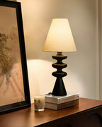 Close view of a black Clérachic table lamp glowing beside a tall leaning frame, with books and a candle on a wooden shelf.