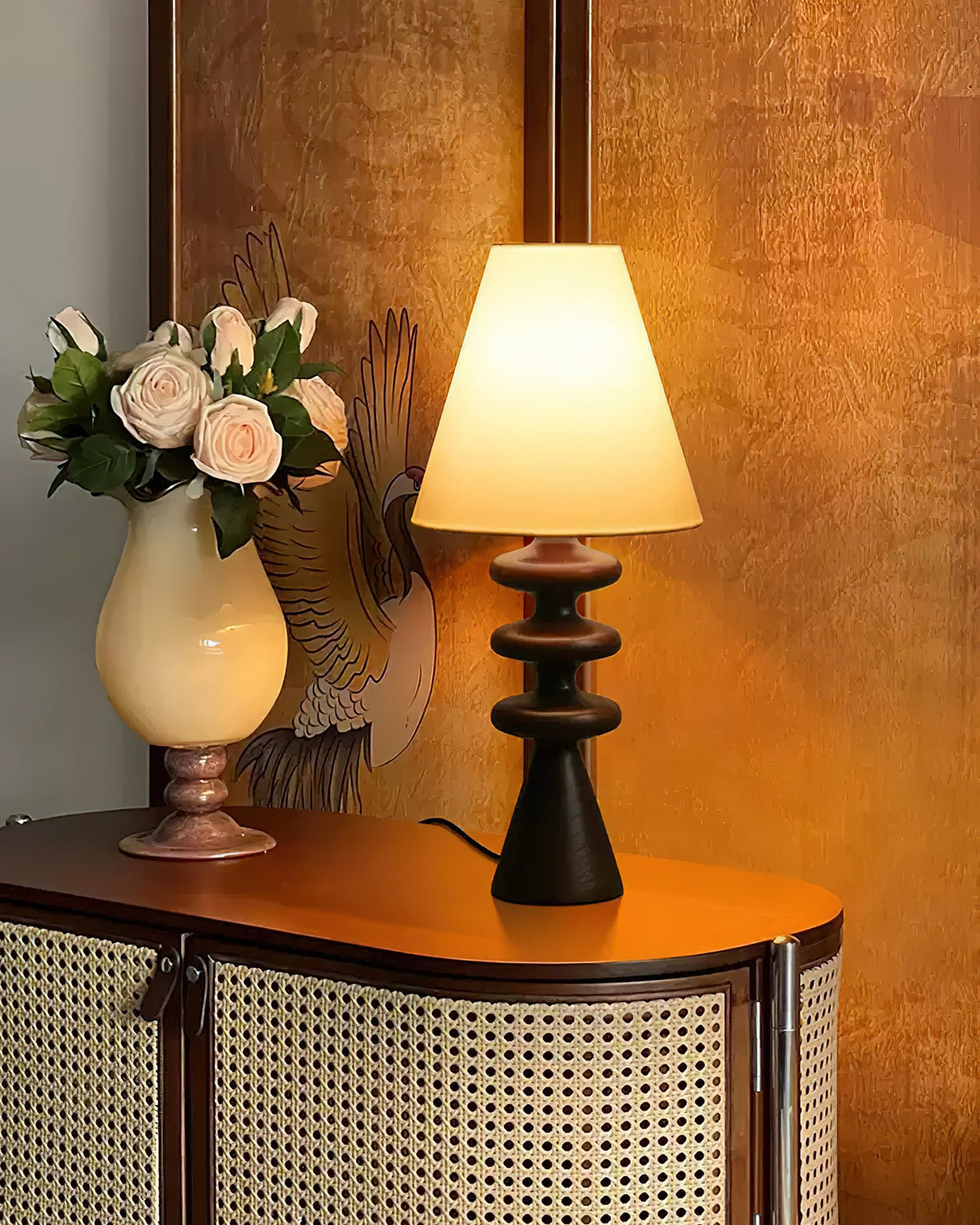 Black Clérachic table lamp lit on a cane-fronted cabinet, a cream vase of roses beside it against a warm amber wall.