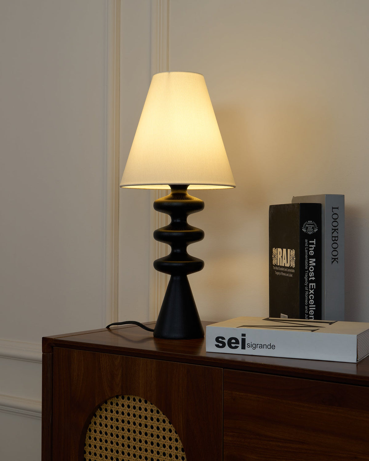 Black Clérachic table lamp switched on atop a walnut sideboard with a round cane panel, books stacked to its right.