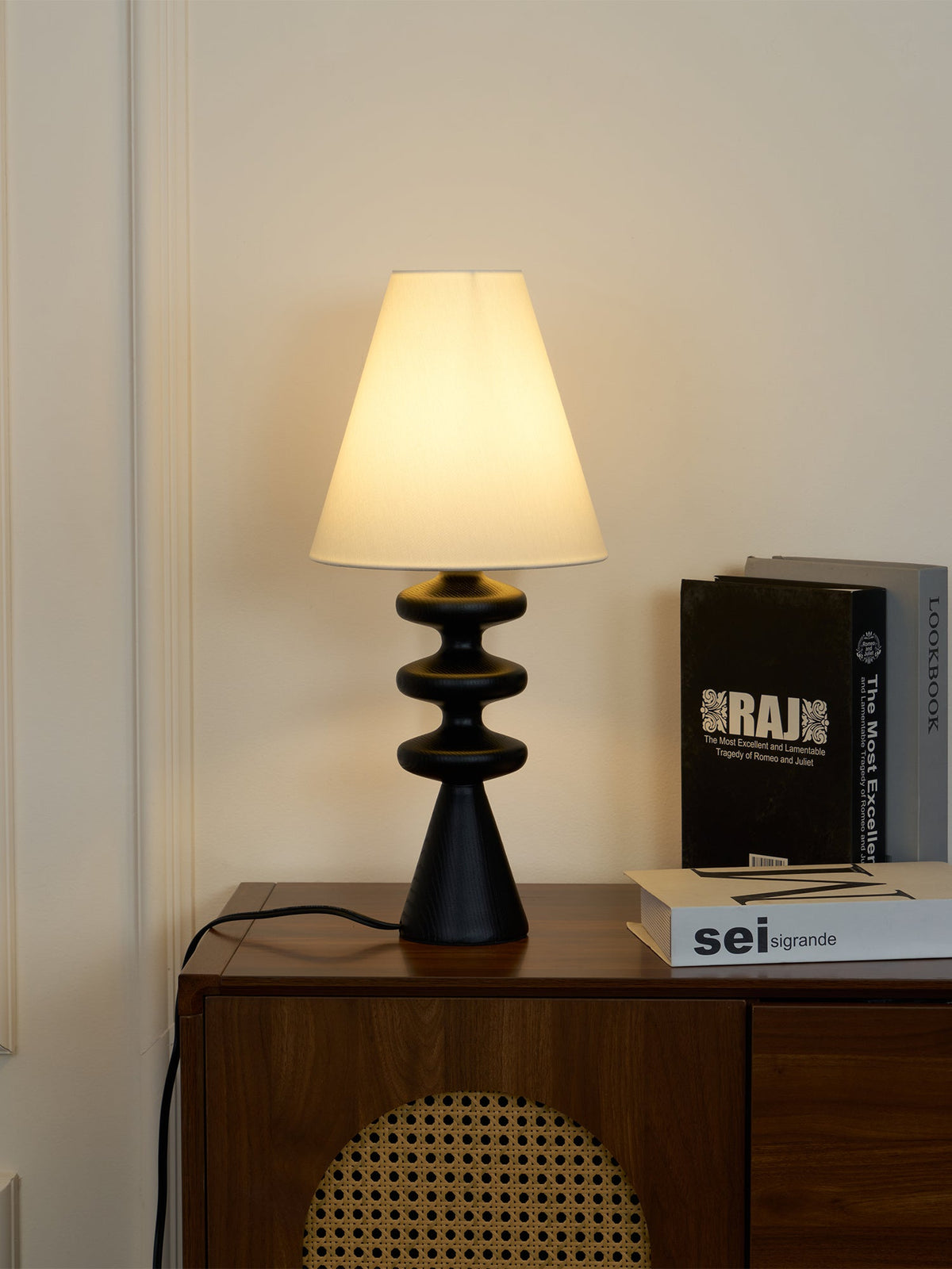 Black Clérachic table lamp at a low glow on a walnut cabinet, the shade lit warm against a panelled white wall.