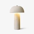 Ceramic table lamp with a domed cream shade, a brass finial and a slim rounded column that glows from within, on a pale backdrop; a Clérachic lamp.