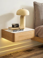 A cream ceramic mushroom-shaped lamp stands on a floating oak bedside shelf with a camera and books, beside a striped pillow; from Clérachic.