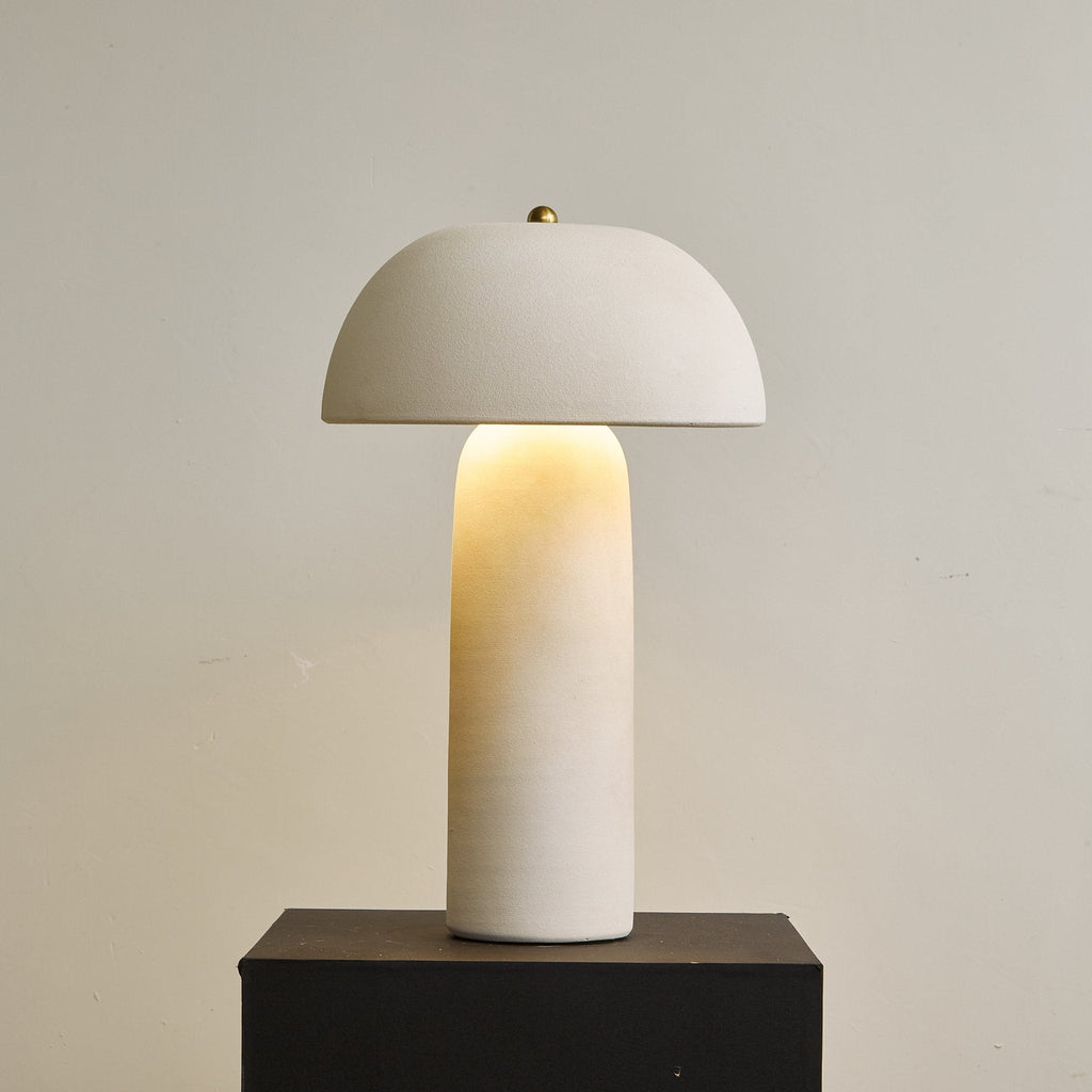 Lit table lamp with a domed cap and a glowing base column on a black plinth, in front of a plain beige wall; a Clérachic design.