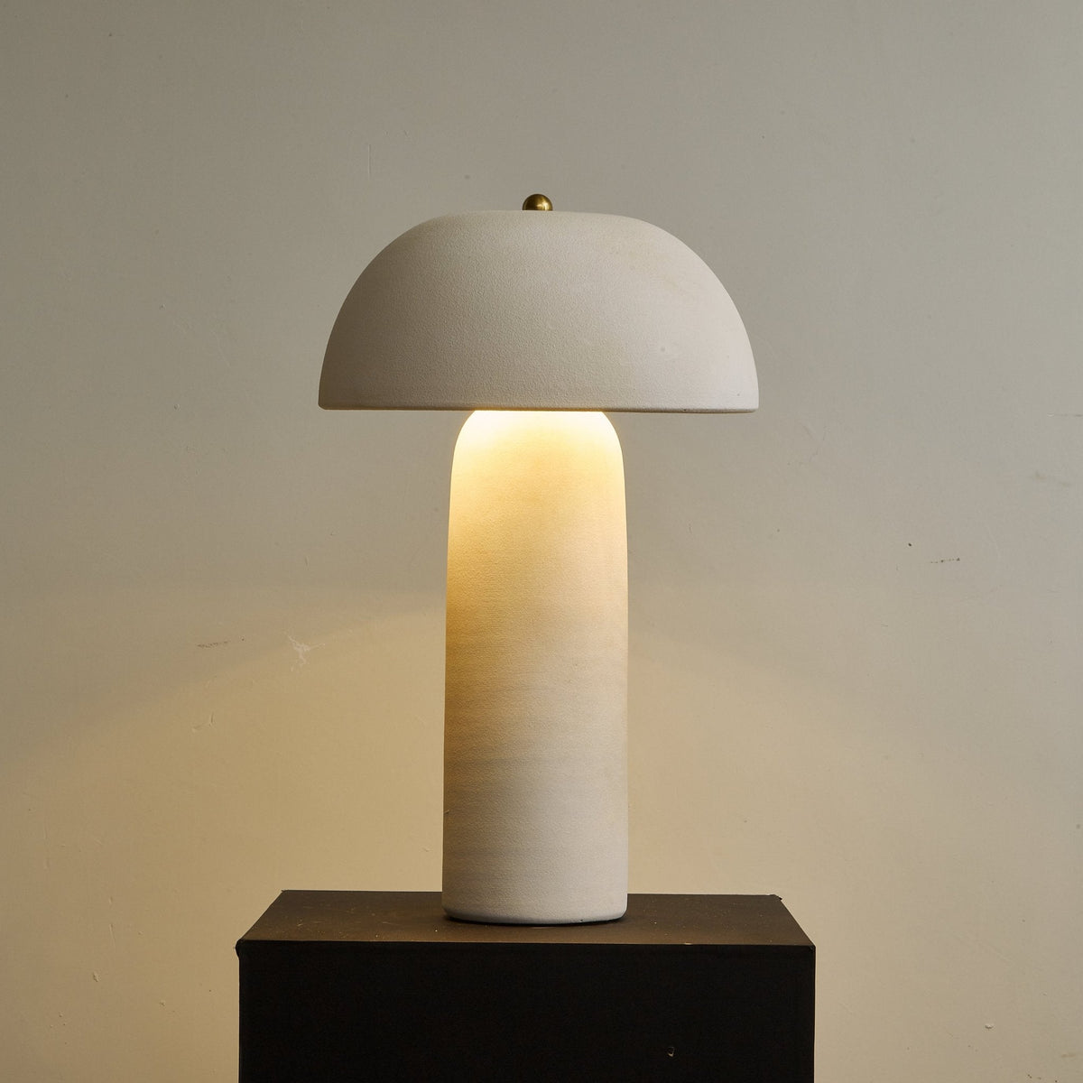 Warm glow from beneath the dome, which lights the matt cream column, on a dark plinth against a taupe wall; a Clérachic table lamp.
