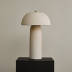 Unlit cream ceramic lamp on a black plinth, showing the rounded dome, the narrow column and a small brass finial; a Clérachic design.