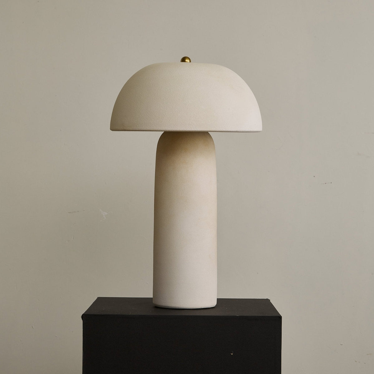 Unlit cream ceramic lamp on a black plinth, showing the rounded dome, the narrow column and a small brass finial; a Clérachic design.