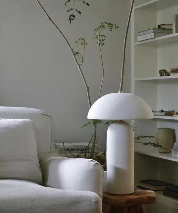 A white version of the lamp sits on a rustic wooden stool beside a linen armchair, a bookcase and a tall branch; from Clérachic.