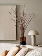 Cream ceramic lamp on a low table below a tall vase of bare branches and a large pale artwork; a Clérachic table lamp.