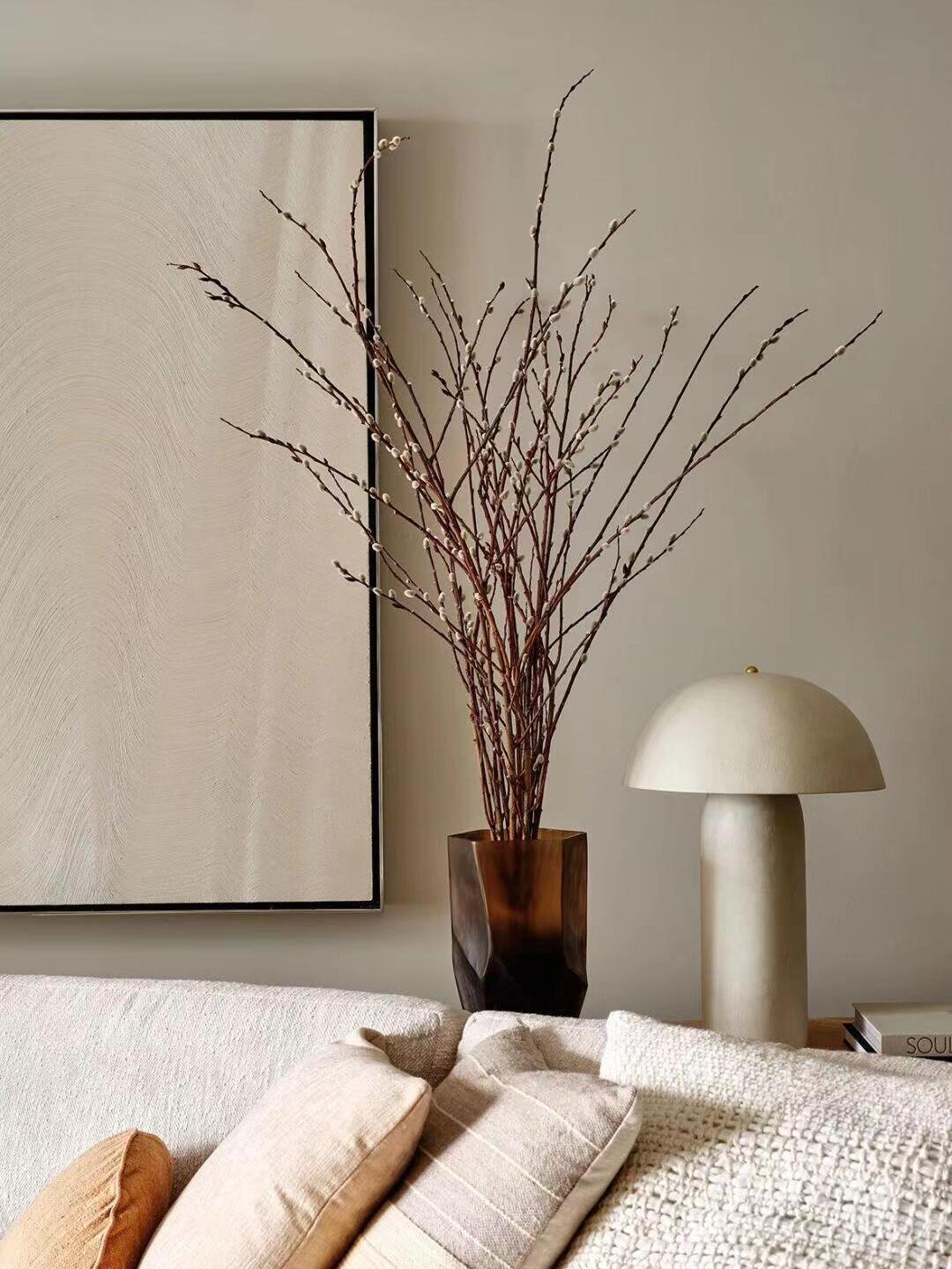 Cream ceramic lamp on a low table below a tall vase of bare branches and a large pale artwork; a Clérachic table lamp.
