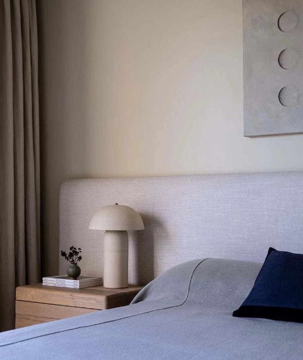 Small cream lamp on a pale wooden bedside table next to a grey-blue bed, with a dark cushion at the edge; this Clérachic lamp.