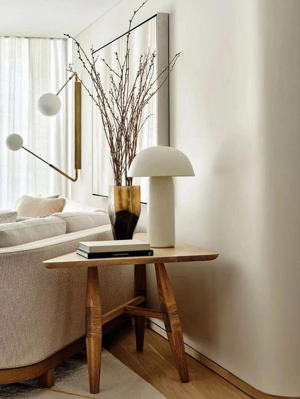 A Clérachic table lamp on a three-legged wooden side table beside a vase of branches and an arm lamp with a white globe.